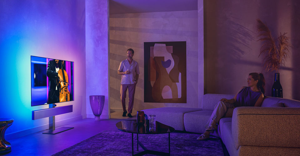 Un homme et une femme dans un salon &eacute;l&eacute;gant regardent un t&eacute;l&eacute;viseur Philips Ambilight diffusant une performance de violoncelle