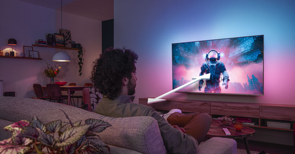 Un homme assis sur un canap&eacute; regarde un t&eacute;l&eacute;viseur Philips Ambilight affichant une sc&egrave;ne d'astronaute color&eacute;e, avec une fl&egrave;che indiquant la distance de visionnage optimale